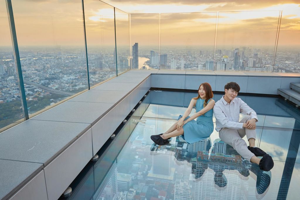 석양이 질 무렵 도시 풍경이 내려다보이는 유리 전망대에 한 젊은 남녀가 앉아 있습니다. 도시 전망이 그 아래에 반영되어 있습니다.