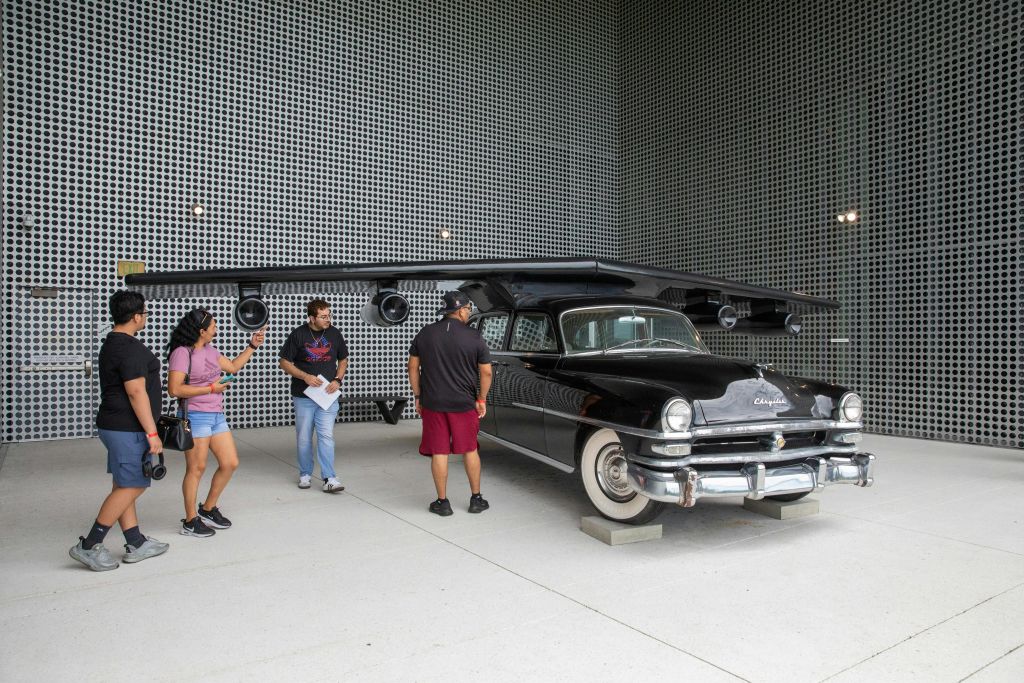 지붕에 날개가 달린 검은색 빈티지 자동차가 특징인 독특한 설치물입니다. 현대적인 환경에서 4명이 관찰할 수 있습니다.