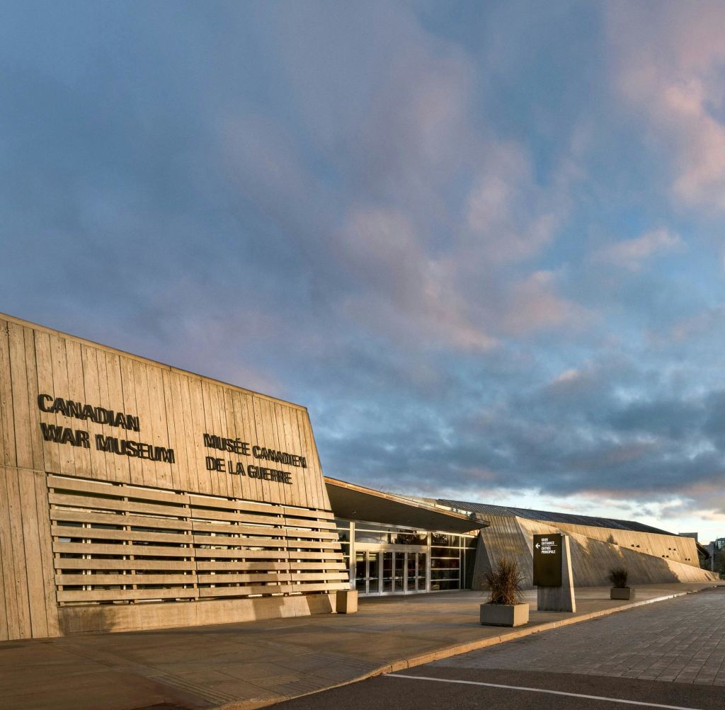 흐린 하늘 아래 "Canadian War Museum" 과 "Musée Canadien de la Guerre" 간판이 있는 현대적인 건물입니다.