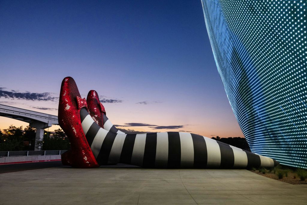 빨간 구두가 달린 줄무늬 다리의 대형 조각상으로, 콘크리트 표면에 옆으로 기울어져 있습니다. 현대적인 건물과 황혼의 하늘을 배경으로.