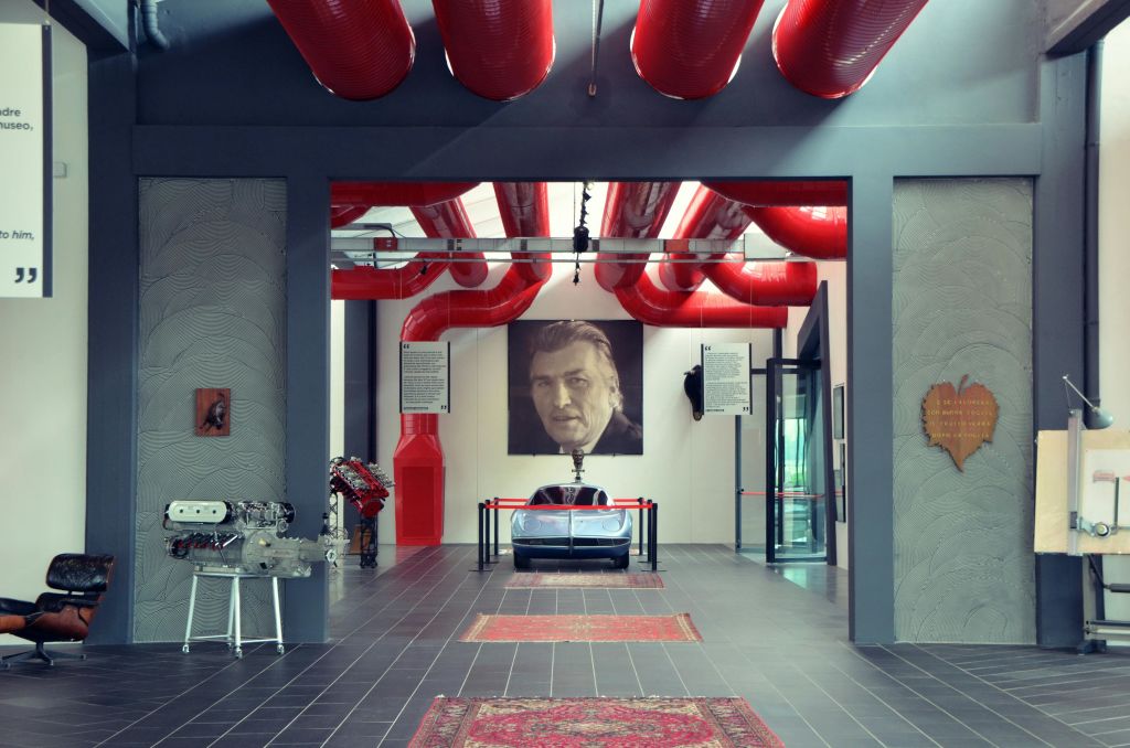 회색 자동차, 빨간색 파이프, 뒷벽에 있는 한 남자의 커다란 초상화가 있는 전시 공간.