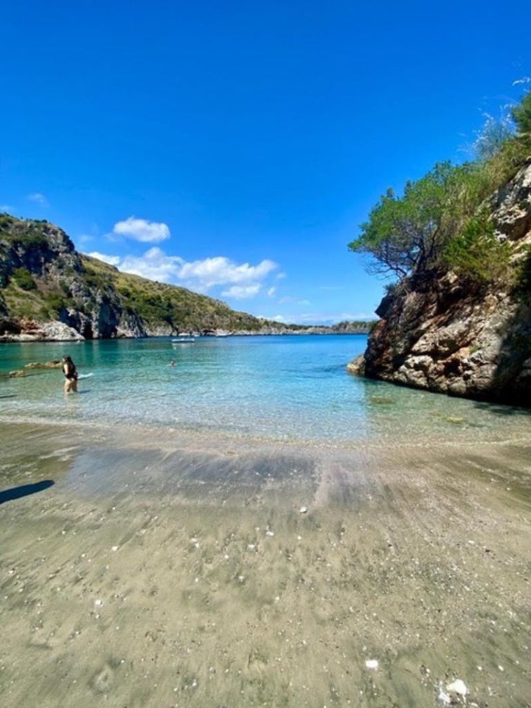 밝고 푸른 하늘 아래 바위 절벽과 나무로 둘러싸인 맑고 푸른 바다가 있는 모래 해변. 얕은 물속을 헤엄치는 사람.