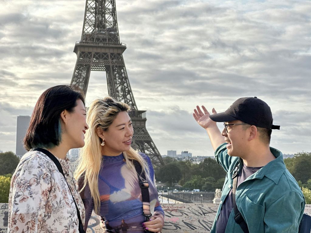 흐린 하늘을 배경으로 에펠탑 근처에서 웃으며 이야기를 나누는 세 사람.
