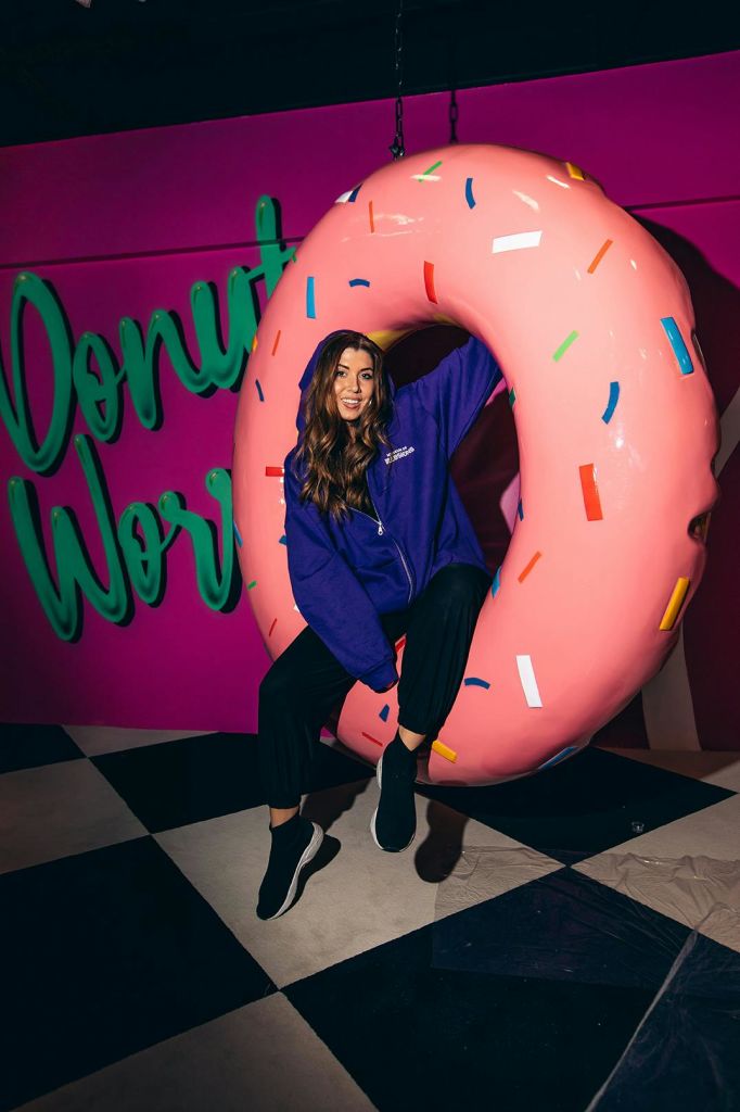 보라색 후디를 입은 여성이 컬러풀한 스프링클이 달린 커다란 분홍색 도넛 안에 앉아 있습니다. 배경 벽에 "도넛 걱정" 이라고 쓰여 있습니다.