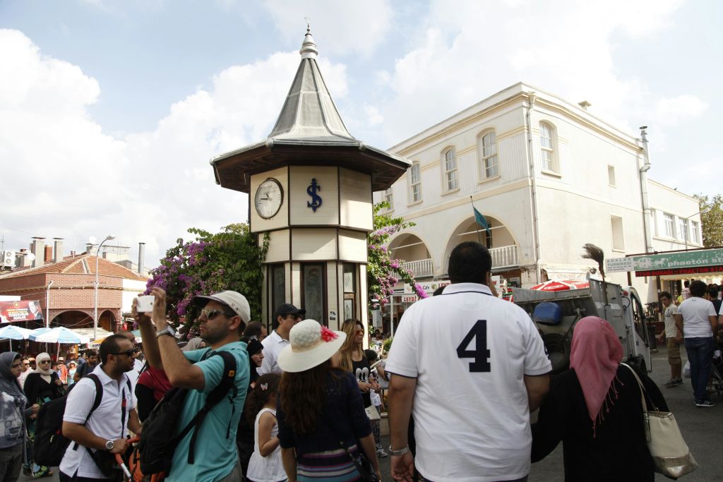 부분적으로 흐린 하늘 아래 파란색 달러 표지판이 있는 작은 시계탑 근처에서 걷고 사진을 찍는 사람들.
