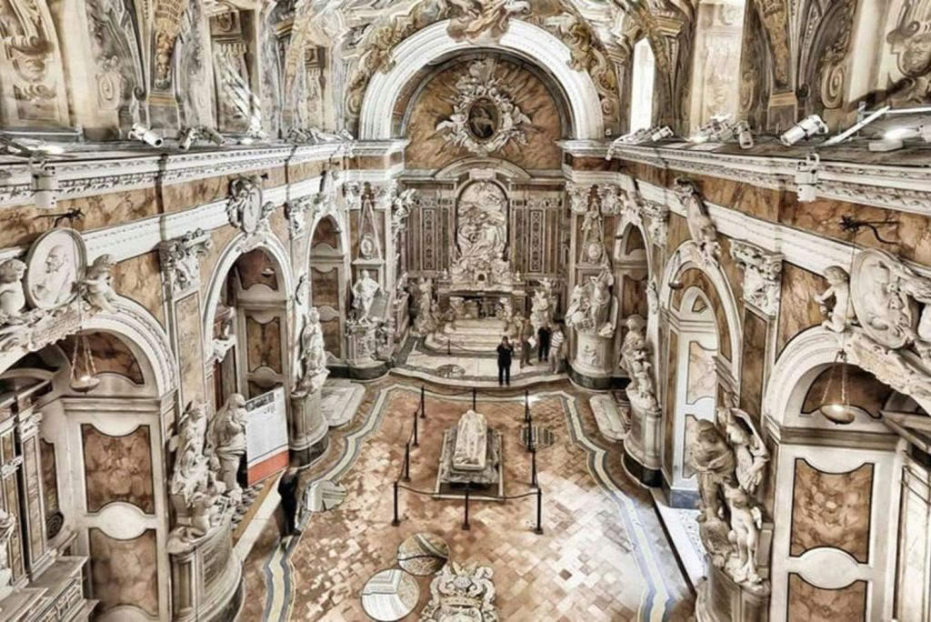 조각품, 아치, 정교한 장식이 있는 화려한 홀로, 중앙의 돌 무덤과 방문객들이 이 공간을 관찰하고 있습니다.