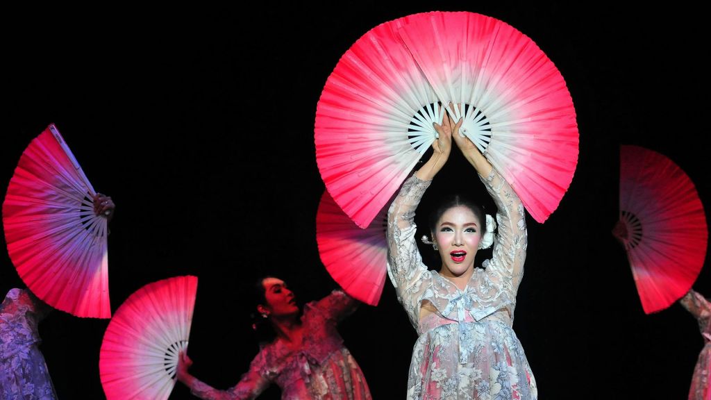 꽃무늬 의상을 입은 연주자가 머리 위로 크고 빨간색과 흰색의 부채를 들고 다른 댄서와 부채를 배경으로 합니다.