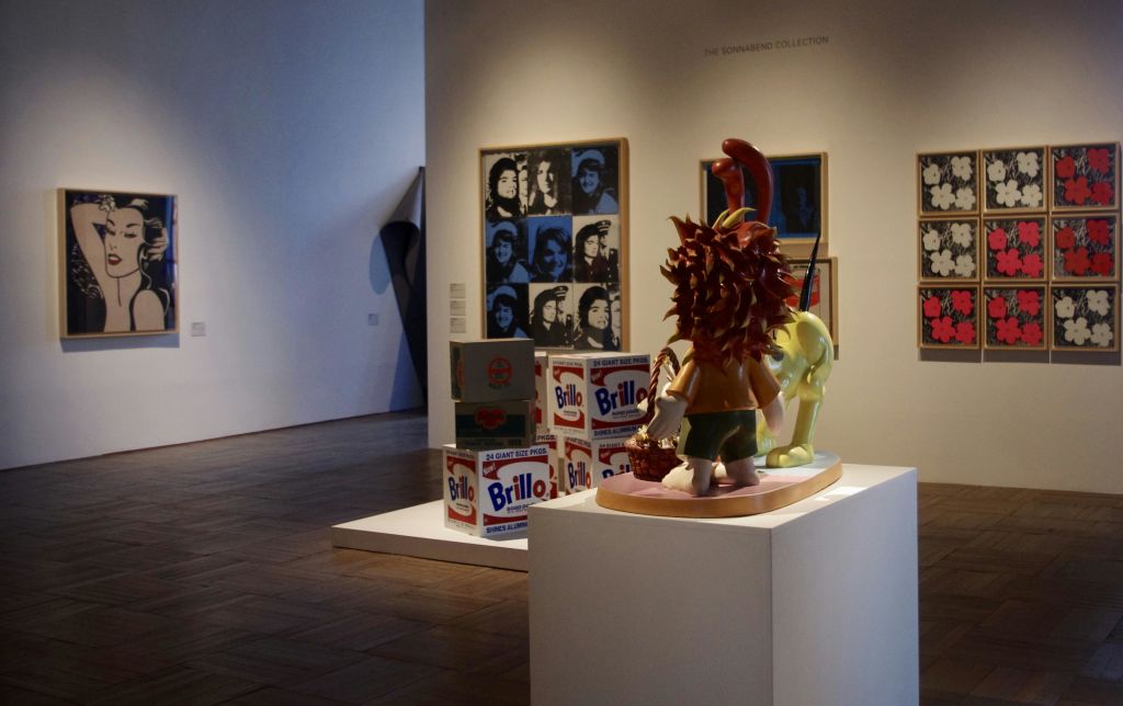 브릴로 상자와 받침대에 있는 만화 조각품, 벽에 액자에 담긴 팝아트 초상화가 있는 아트 갤러리 장면.