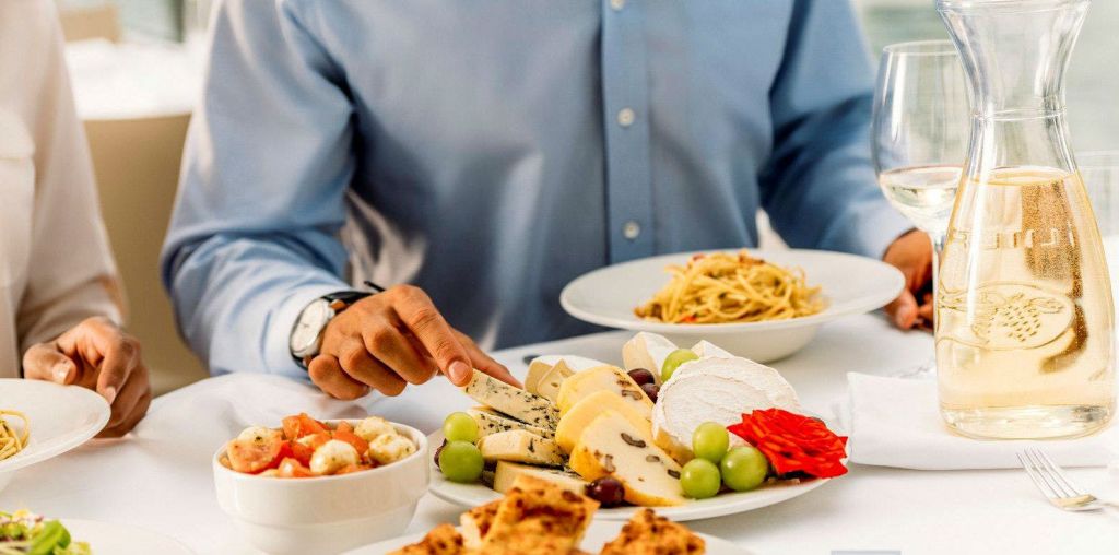 다양한 치즈, 포도, 붉은 장식이 있는 접시에서 치즈를 선택하는 파란색 셔츠를 입은 사람. 배경에는 파스타 요리와 빵이 있습니다.