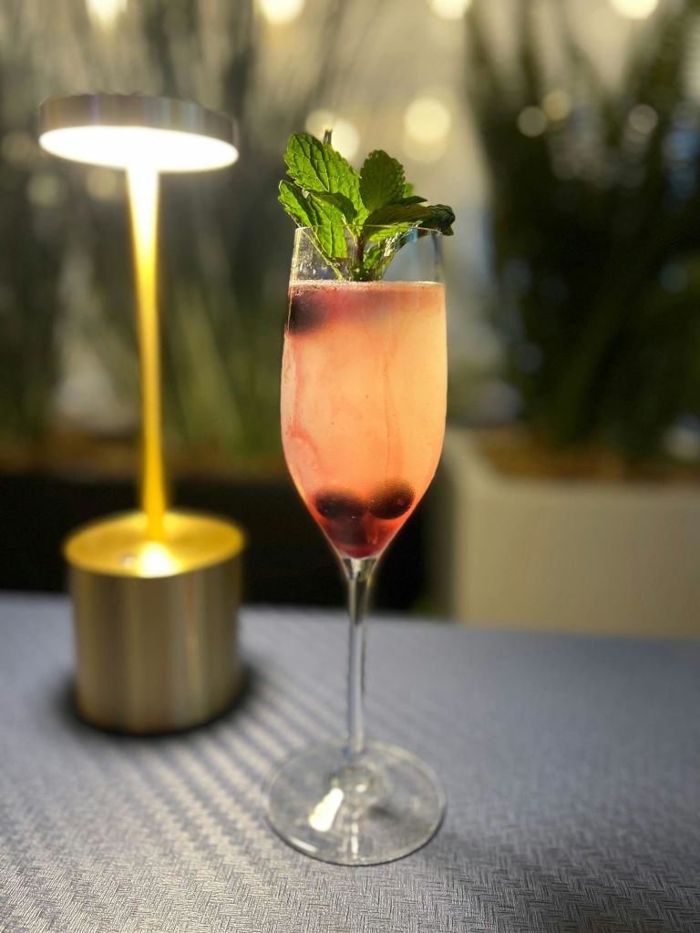 조명 램프 옆 테이블에 민트 잎과 열매로 장식된 핑크색 음료를 곁들인 샴페인 플루트입니다.