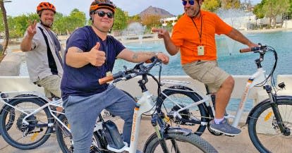 주황색 헬멧을 쓴 두 사람이 공원의 분수대 근처에서 자전거를 들고 서 있는데, 한 명은 엄지손가락을 치켜세우고 다른 한 명은 평화 표시를 하고 있습니다.