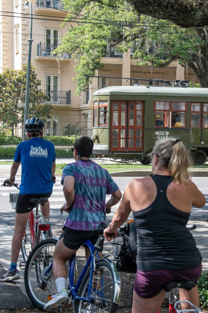 화창한 날 녹색 노면 전차 근처의 교차로에서 자전거를 타는 사람 세 명이 잠시 멈추며, 주거용 건물과 나무를 배경으로 이야기를 나눕니다.