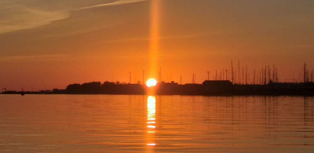실루엣의 건물과 돛대의 지평선 뒤에서 태양과 함께 잔잔한 물 위로 석양을 감상하며 수직적인 빛 반사를 연출합니다.