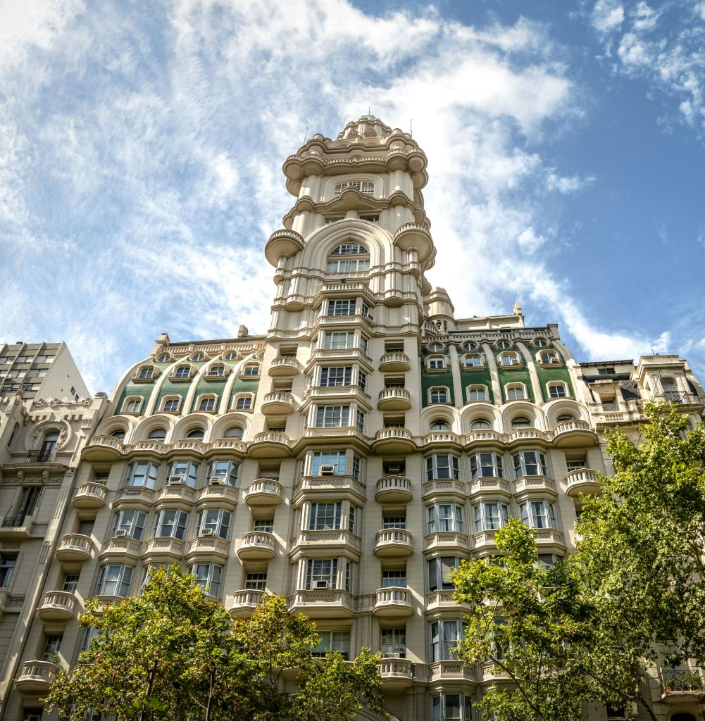 아치형 창문과 장식 디테일이 돋보이는 높고 화려한 건물로, 구름이 있는 푸른 하늘을 배경으로 나무로 둘러싸여 있습니다.