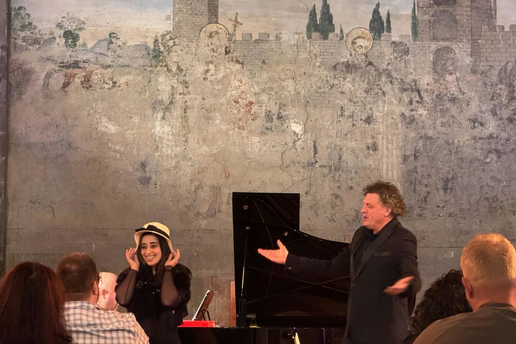 모자를 쓴 여성이 피아노와 오래된 벽화를 배경으로 공연 도중 미소를 짓고 제스처를 취하고 있습니다.