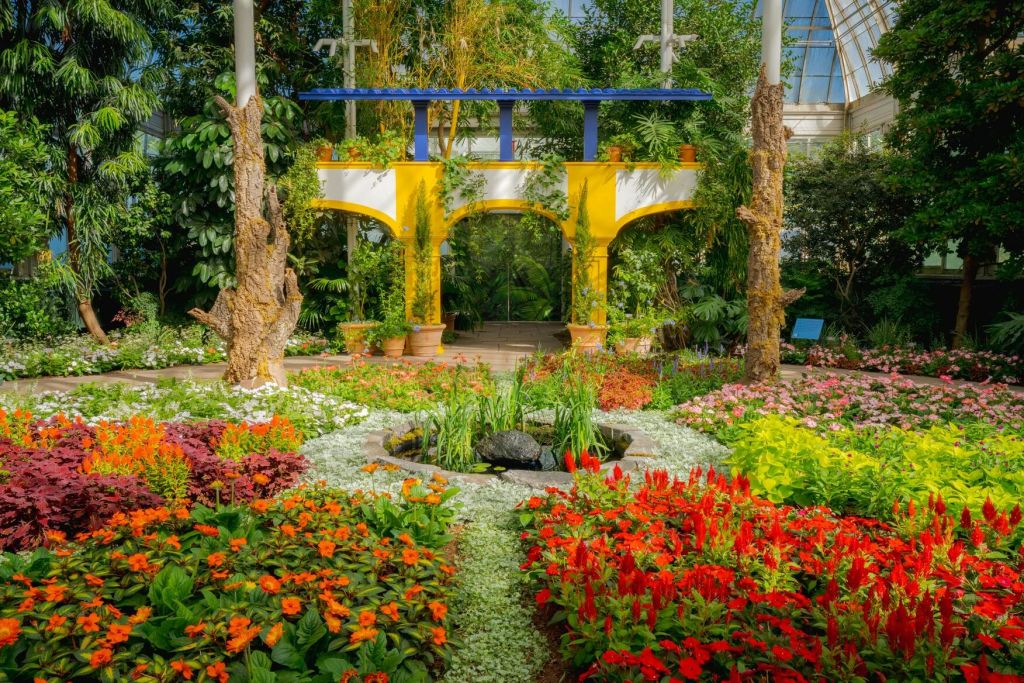 다양한 꽃과 화분, 작은 연못, 유리 천장 아래 노란색과 파란색 아치형 통로가 있는 활기찬 정원입니다.