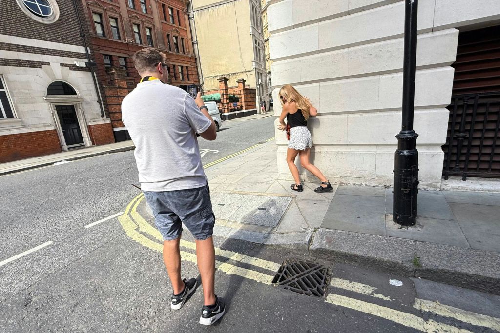 대낮에 도시의 거리에서 건물 모퉁이를 배경으로 포즈를 취하는 여성을 촬영한 남성.