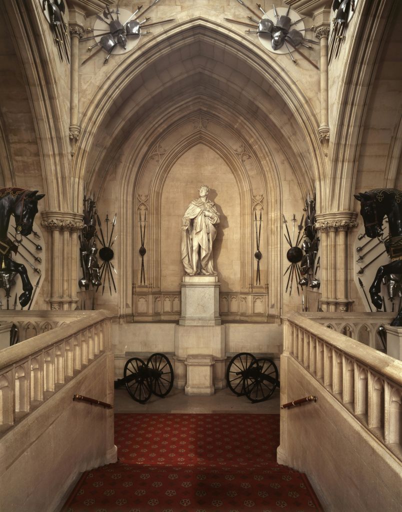 중앙에 동상이 있는 고딕 양식의 홀, 갑옷, 벽에 걸린 무기, 아래에 있는 대포. 레드 카펫이 깔린 계단이 내려갑니다.