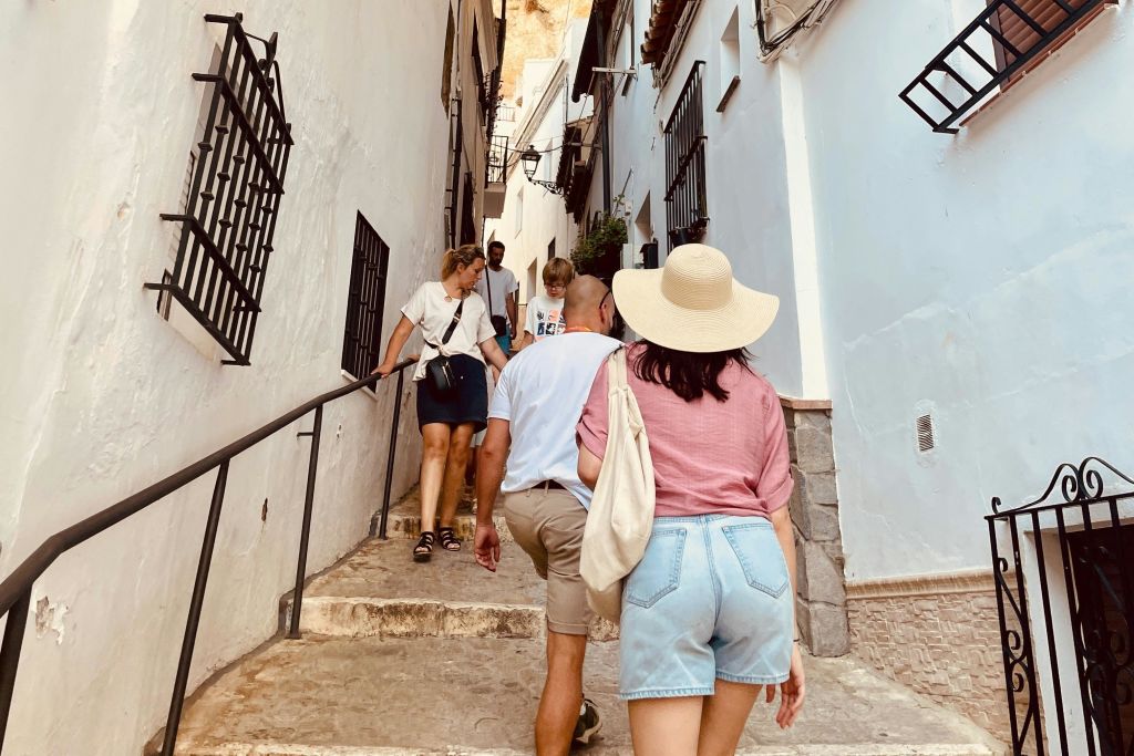 화창한 날 하얀 건물과 철제 창문 그릴이 있는 좁은 골목을 걷는 사람들.