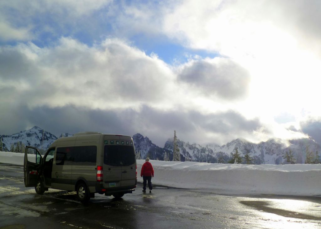 붉은 재킷을 입은 사람이 눈 덮인 산과 흐린 하늘을 배경으로 젖은 도로의 주차된 밴 근처에 서 있습니다.