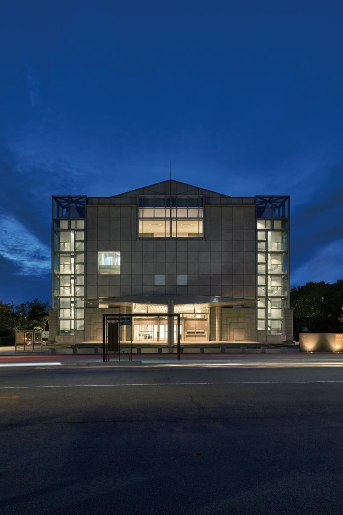 커다란 유리 섹션이 있는 현대적인 입방 건물이 밤에는 깊고 푸른 하늘을 배경으로 조명을 받습니다.