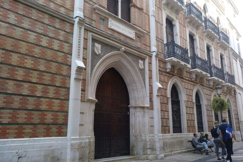 "Hospital de S. Tomas" 라고 표시된 아치형 나무 문이 있는 벽돌 건물로, 두 개의 창문과 근처에 앉아 있는 사람이 옆에 있습니다.
