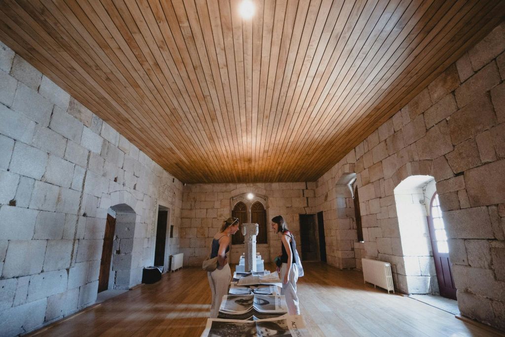 돌담, 나무 천장, 아치형 출입구가 있는 방에서 두 여성이 유물을 살펴보고 있습니다. 신문이 테이블 위에 있습니다.