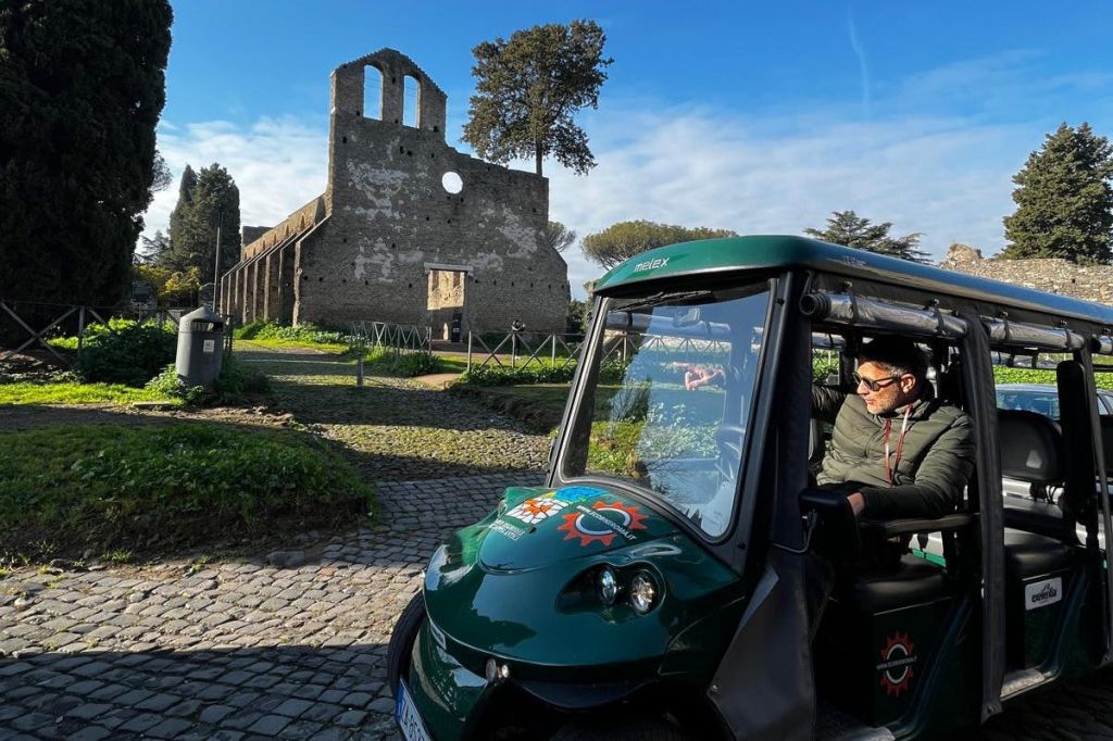 고대 유적과 나무를 배경으로 자갈길에 주차된 녹색 투어 카트 안에 한 남성이 앉아 있습니다.