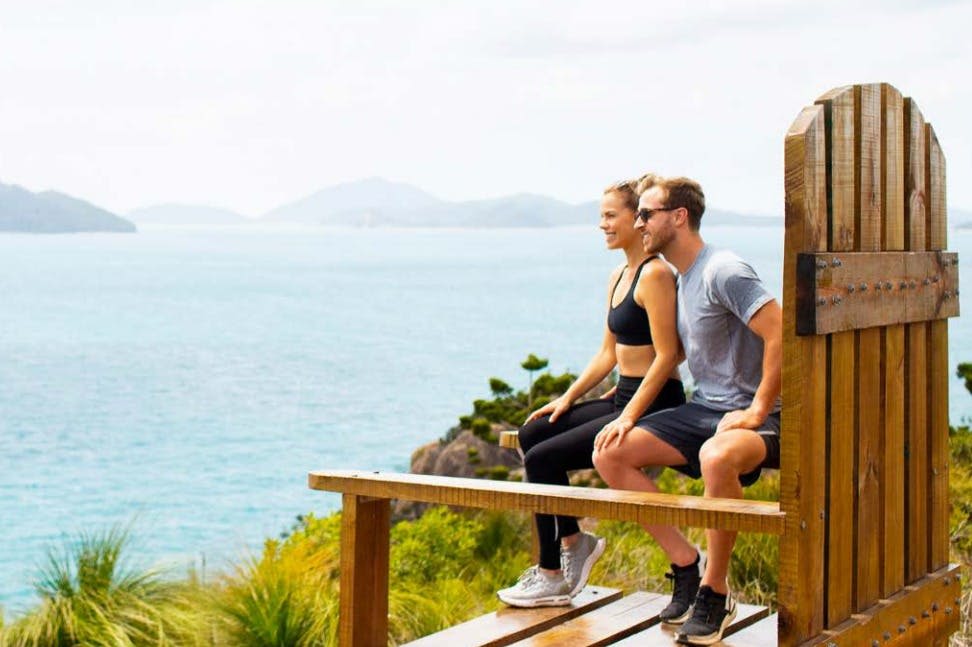 부부가 나무 벤치에 앉아 산이 내려다보이는 아름다운 해안 전망을 감상하고 있습니다.