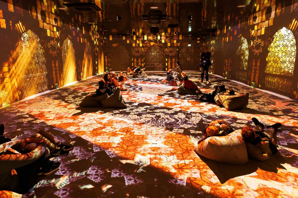 사람들은 바닥과 벽에 화려하고 정교한 조명으로 비춰진 방에서 콩 주머니에 앉아 있습니다.