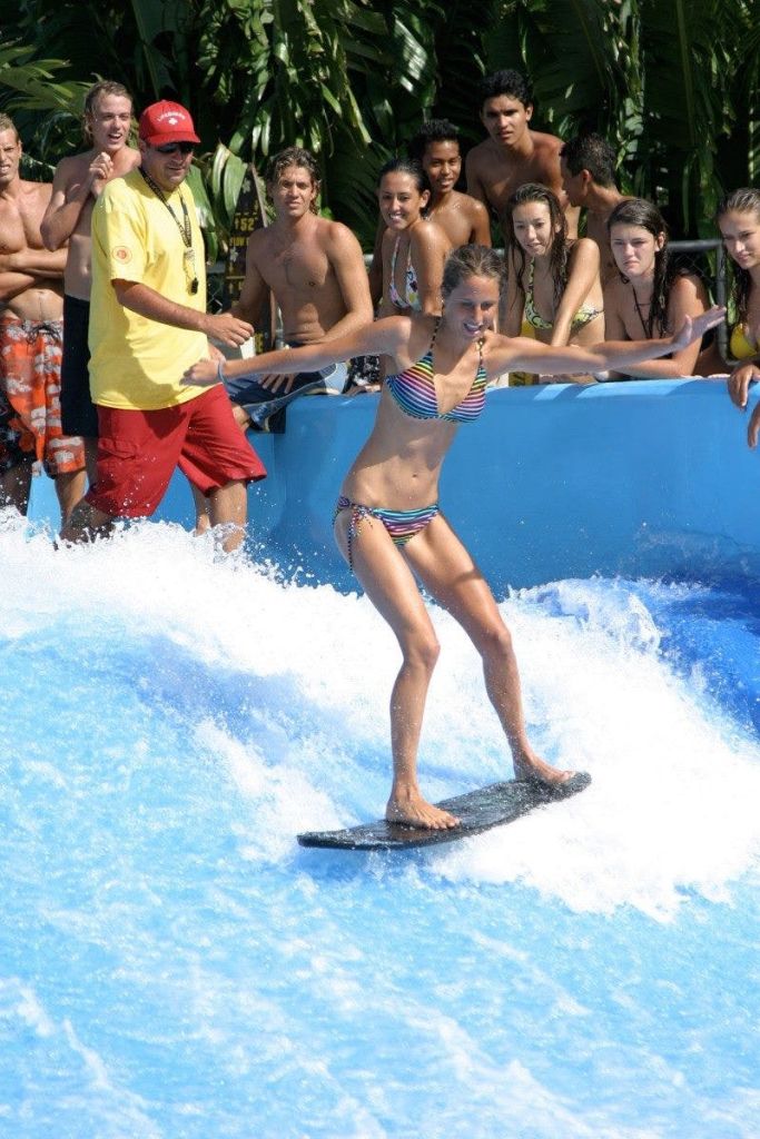 한 여성이 시뮬레이션 웨이브에서 균형을 잡는 동안 한 무리의 사람들이 수영장 가장자리에서 지켜보고 있습니다.