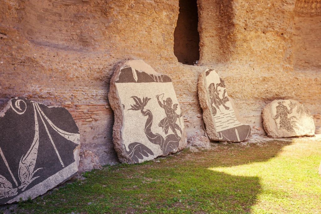 신화 속 인물이 그려진 고대 석조 모자이크는 잔디가 우거진 지역의 풍화된 벽돌 벽에 기대어 있습니다.