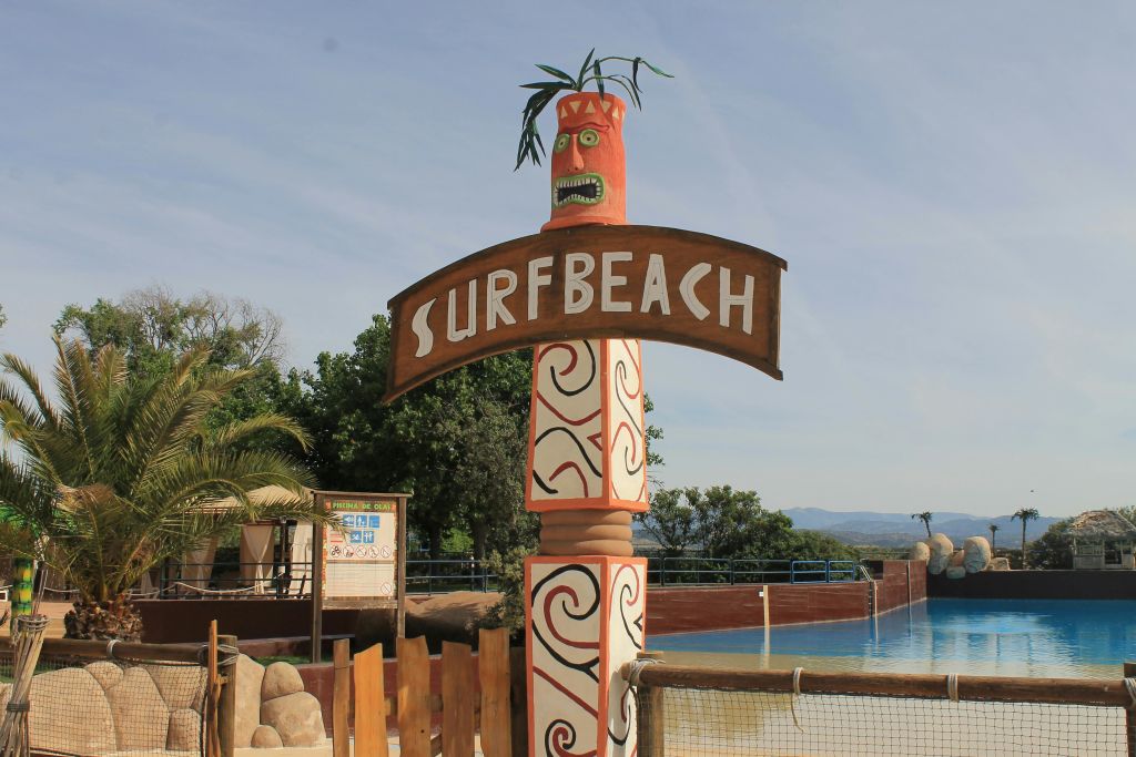 "서핑 비치" 텍스트가 있는 표지판, 티키 얼굴로 장식되어 있으며, 수영장 근처에 있으며 나무와 맑은 하늘로 둘러싸여 있습니다.