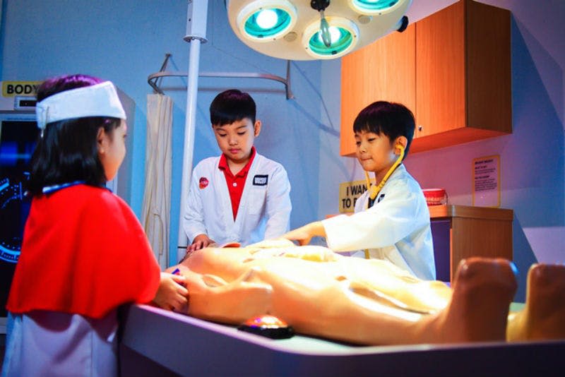 의사 복장을 한 세 명의 어린이가 수술 조명 아래에서 더미에 대한 수술을 수행합니다.