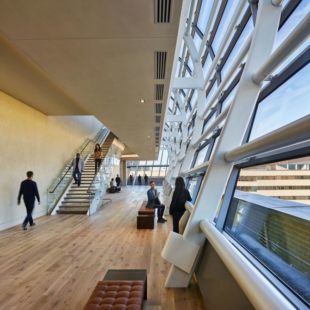 커다란 창문, 목재 바닥, 계단이 있는 햇살이 내리쬐는 모던한 인테리어에서 사람들이 걷고 앉아 있습니다.