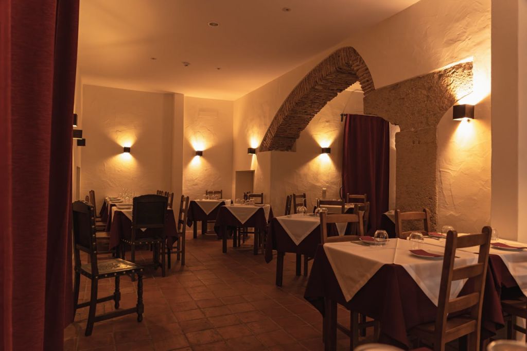 탁자, 나무 의자, 벽면이 비치는 아치형 출입구 위에 흰색 식탁보가 놓인 어두운 조명의 레스토랑입니다.