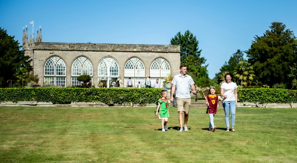 화창한 날에 아치형 창문과 녹음이 우거진 건물 앞 잔디밭을 따라 4인 가족이 산책합니다.