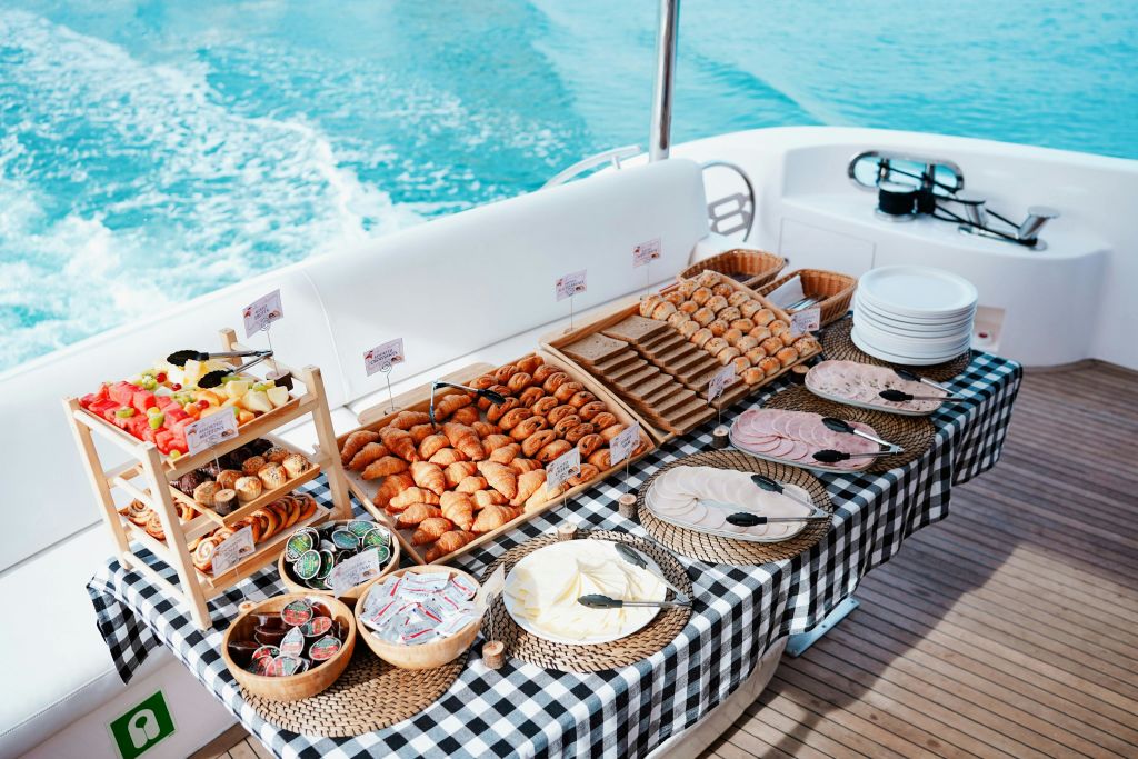 크루아상, 콜드컷, 과일, 다양한 양념을 바닷가 체크무늬 식탁보에 담은 보트 조식 뷔페.