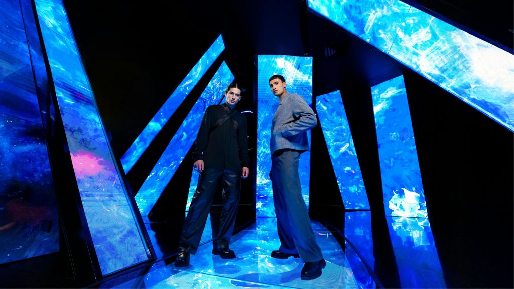 커다란 파란색 조명 기하학적 패널이 있는 미래 지향적인 분위기에서 두 남자가 포즈를 취하고 있습니다. 어둡고 현대적인 옷을 입고 있습니다.