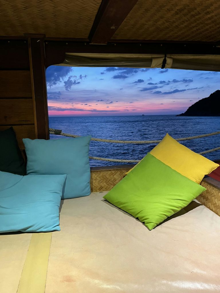 바다를 조망할 수 있는 아늑한 좌석 공간에 밝은 색상의 베개가 배치되어 있으며 나무 프레임으로 일몰을 감상할 수 있습니다.