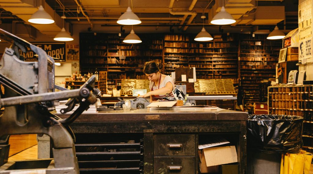 책 선반과 다양한 도구로 가득 찬 조명이 희미한 작업장의 어수선한 책상에서 일하는 사람.