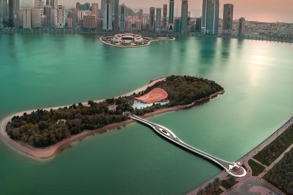 현대적인 돔 지붕 건물이 있는 섬으로, 청록색 바다 위로 다리가 연결되어 있으며, 도시의 스카이라인이 배경으로 펼쳐집니다.