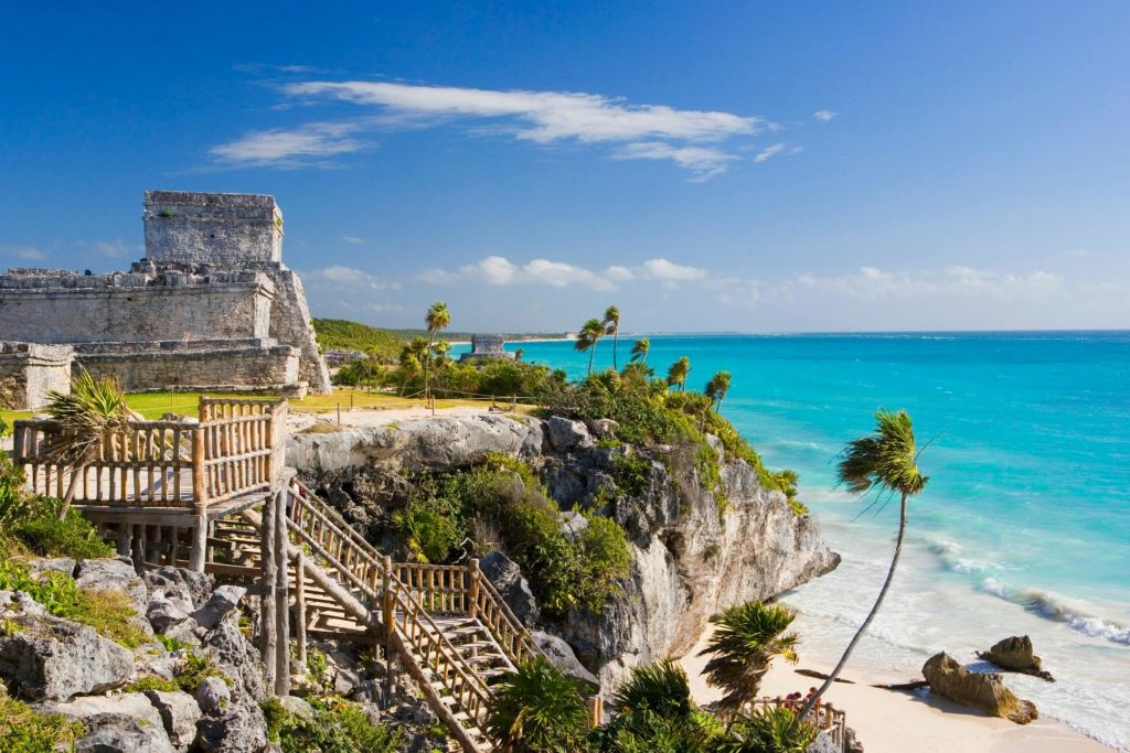 청록색 해변이 내려다보이는 절벽의 석조 유적지와 나무 계단, 야자수와 부분적으로 흐린 푸른 하늘.