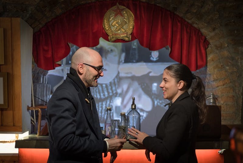 어두운 복장을 한 두 사람이 빈티지 장식과 붉은 커튼에 새겨진 소련 엠블럼이 있는 바에서 대화를 나누고 있습니다.