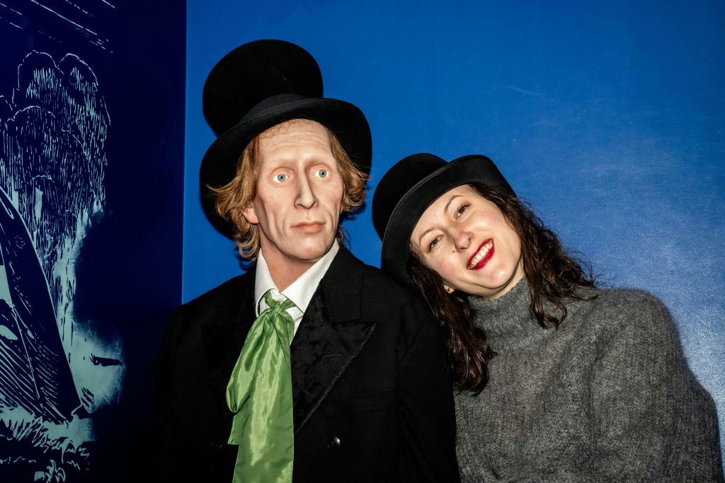 회색 스웨터를 입은 여성이 양복을 입은 남성의 왁스 피규어와 파란색 벽에 초록색 갈매기가 달린 모자 옆에서 몸을 기울이고 미소를 짓고 있습니다.