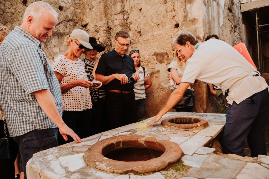 한 무리의 사람들이 석조 테이블에 원형의 함몰이 있는 역사적인 석조 구조물에서 투어 가이드의 말을 듣고 있습니다.