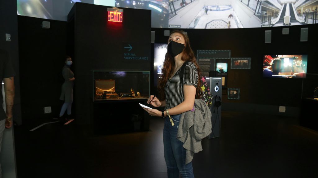 줄무늬 셔츠와 검은색 마스크를 착용한 여성이 전시 케이스와 '출구' 표지판 근처의 박물관 전시 공간에서 메모를 하고 있습니다.