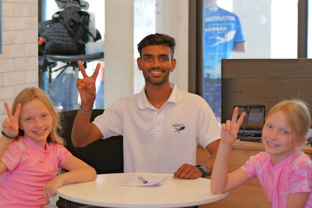 흰색 폴로 셔츠를 입은 한 남성이 평화 표지판을 들고 있는 테이블에 앉아 있으며, 앞쪽에 두 명의 어린이가 부분적으로 보입니다.