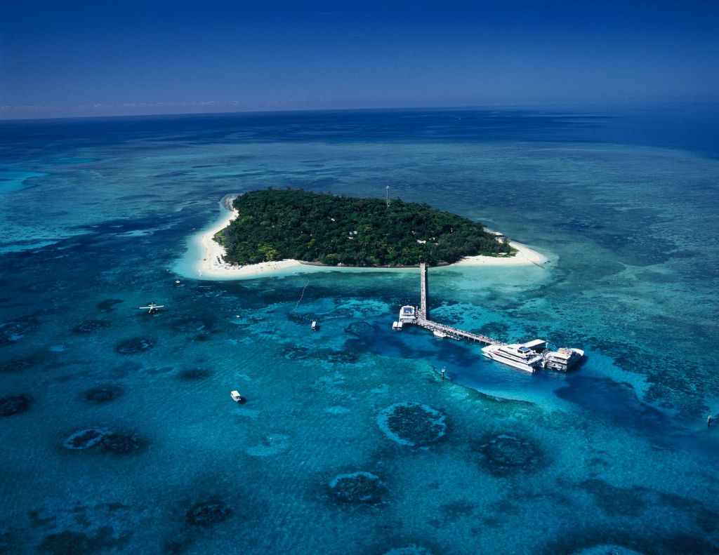 하얀 모래 해변과 맑고 푸른 물로 둘러싸인 녹음이 우거진 작은 열대 섬의 공중 전망을 감상할 수 있는 보트가 정박해 있습니다.