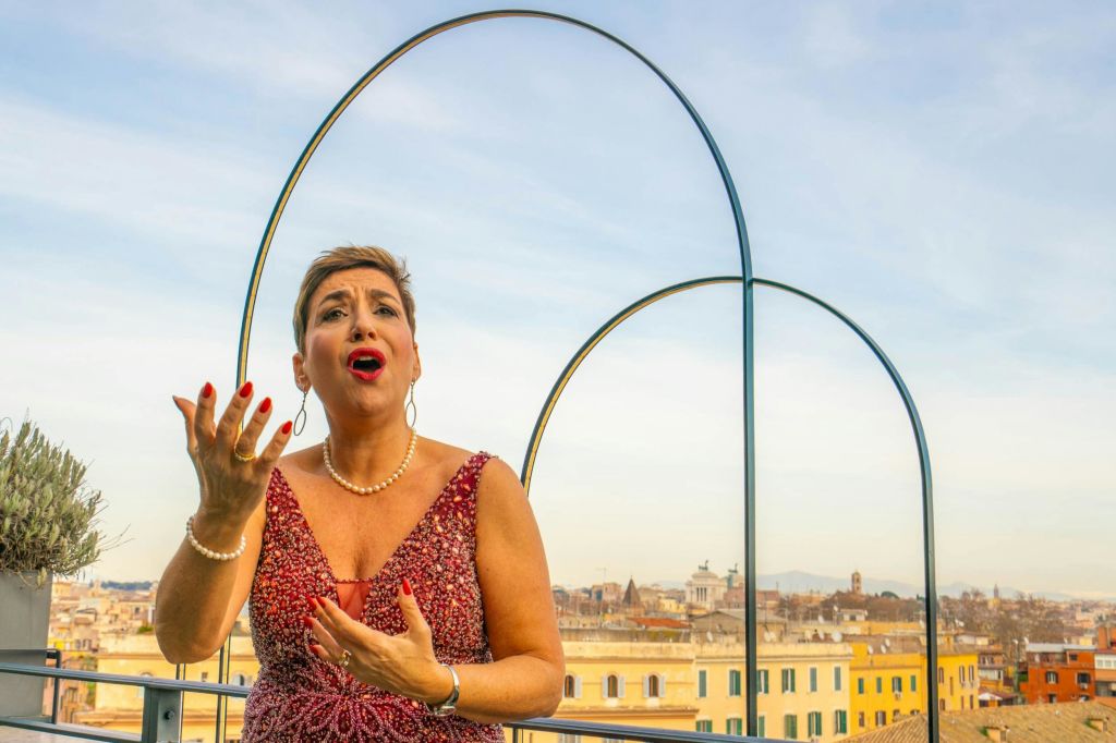 도시 풍경과 푸른 하늘을 배경으로 옥상 테라스에서 빨간 드레스를 입은 사람이 열정적으로 노래하고 있습니다.